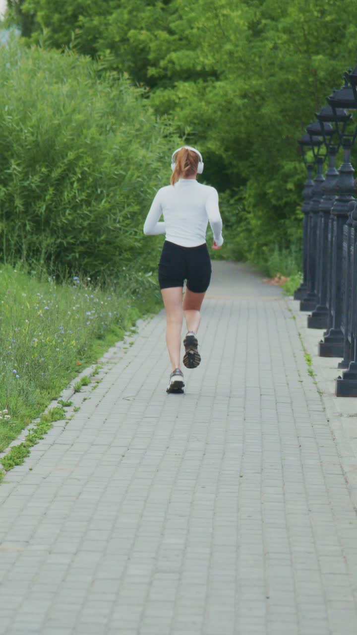 A Young Woman Jogging Along a Scenic Pathway, Enjoying Her Run While Listening to Music on Headphones, Surrounded by Lush Greenery and a Relaxing Atmosphere