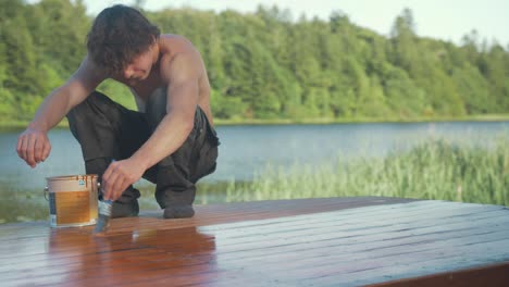 young carpenter sits in primitive squat painting roof planking of wooden boat with exterior protective paint