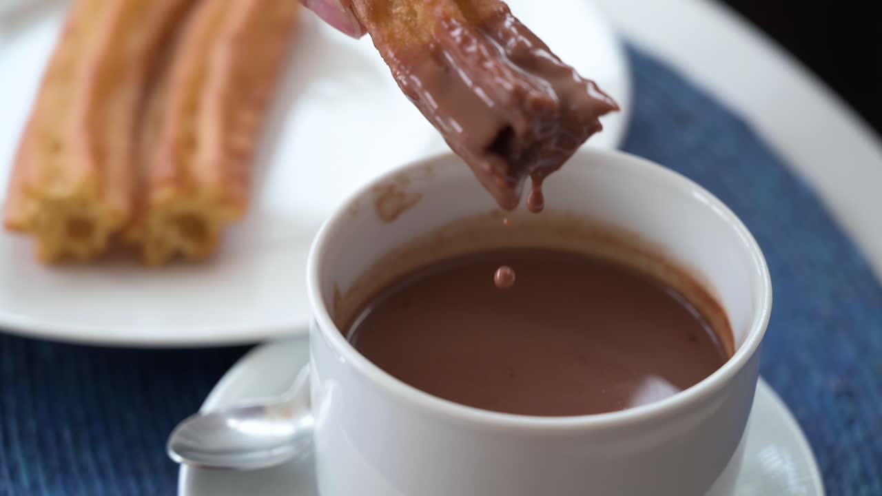 un metraje en cámara lenta, una mano sumerge un churro español en salsa de chocolate caliente