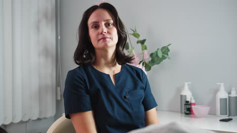 portrait view of aesthetician holding gua sha tool and sighing with relief while seated confidently and smiling after completing facial treatment in spa room with elegant floral decor