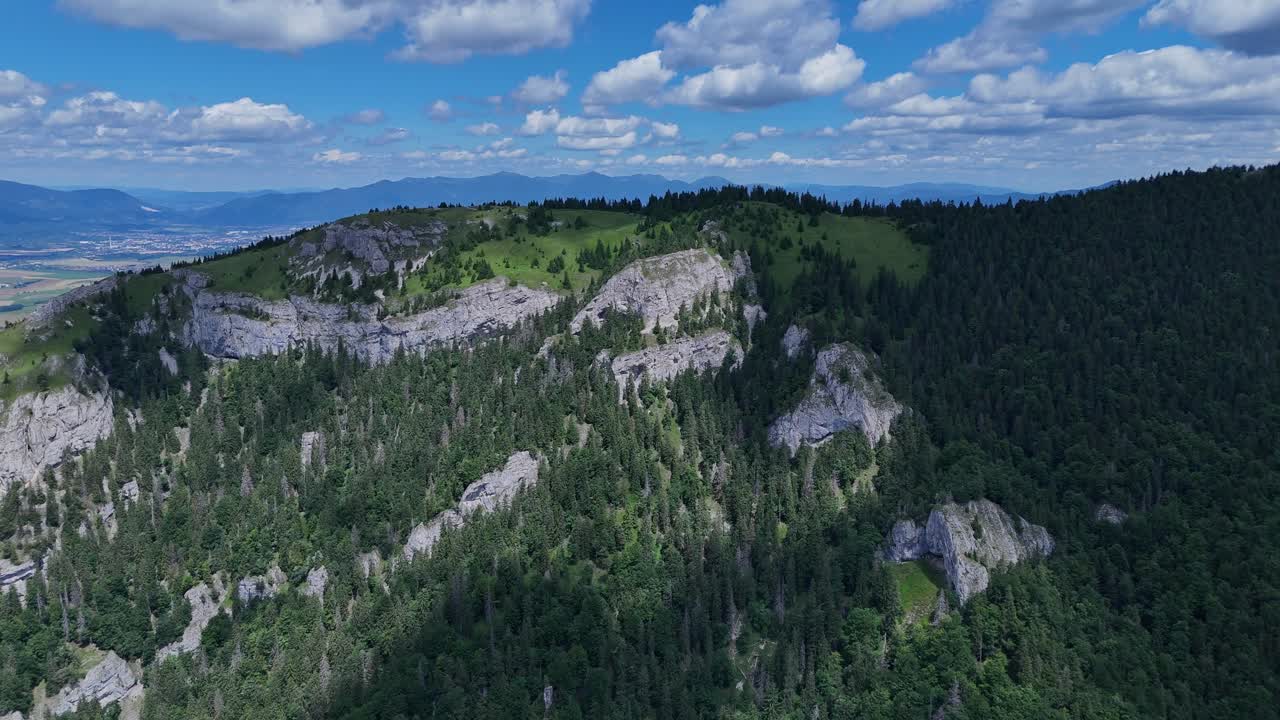 Aerial drone footage reveals a mountain range beyond rugged rocky slopes and dense green forests in the foreground, capturing the natural beauty of Slovakia’s alpine landscape. Location: Slovakia
