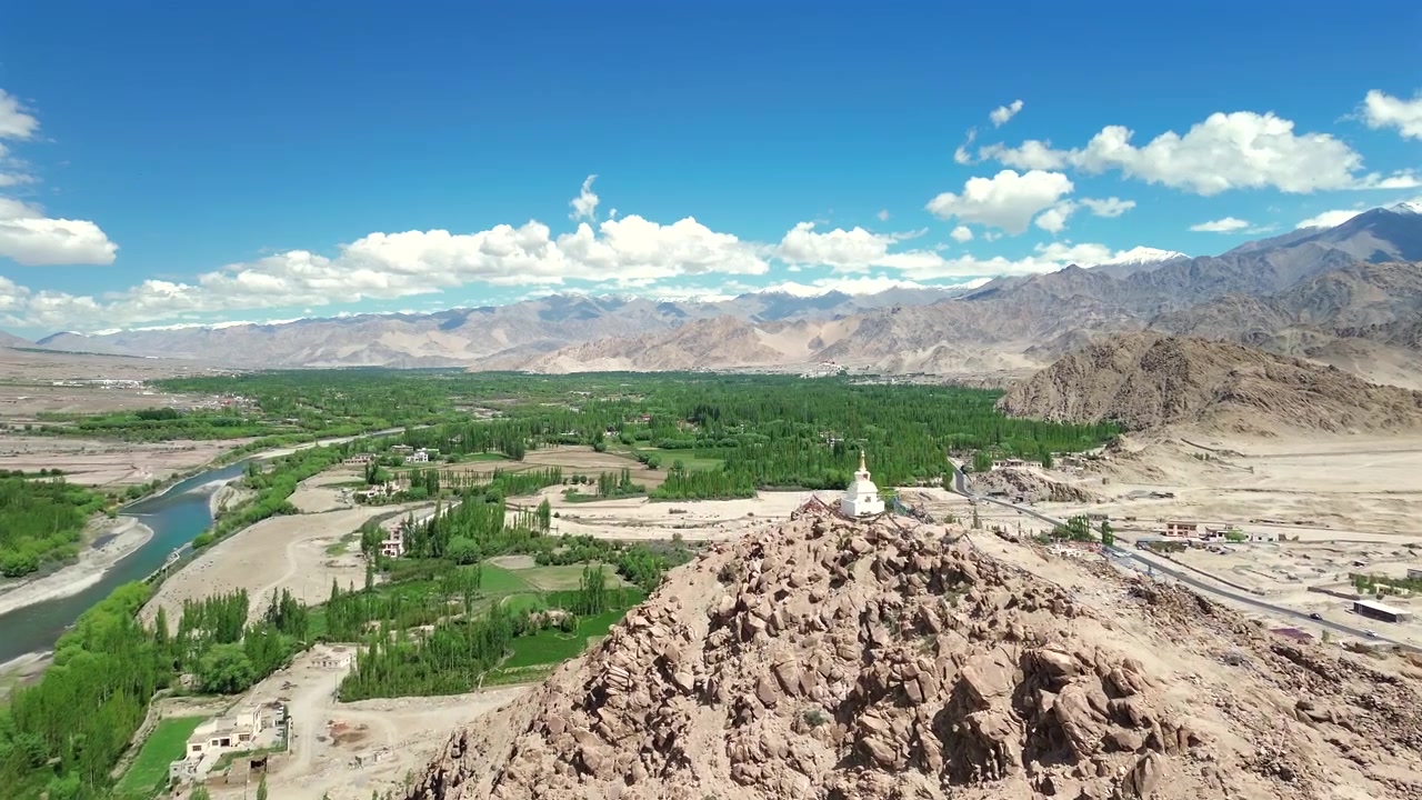 Højvinkel droneoptagelse, der sporer og stiger op over en barsk klippekam for at afsløre en hvid buddhistisk chorten stupa med udsigt over den frodige grønne Indus-floddal i Leh Ladakh, Indien
