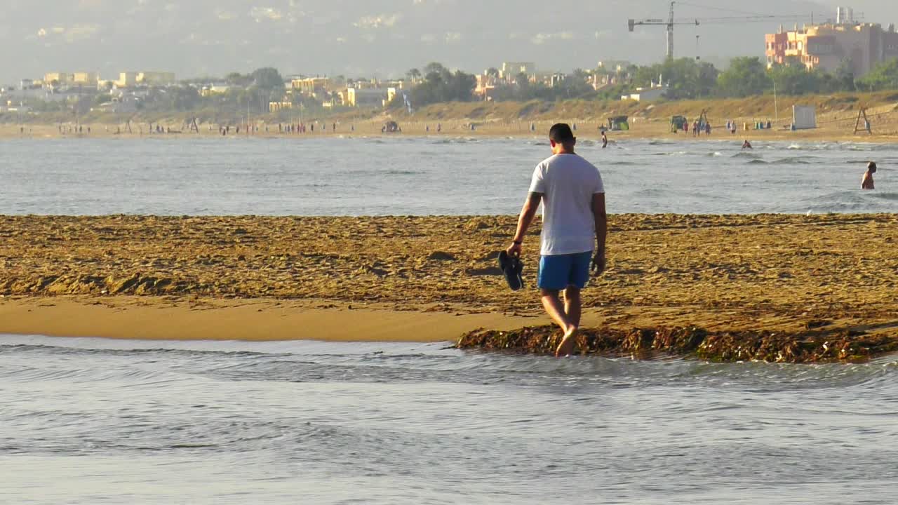 joven caminando en olas en la playa de arena en españa, tiro ancho de cámara lenta