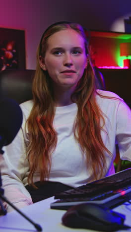 young woman with long blonde hair smiling at the camera while sitting at a desk in front of a microphone, keyboard, and mouse. she appears to be streaming or gaming.