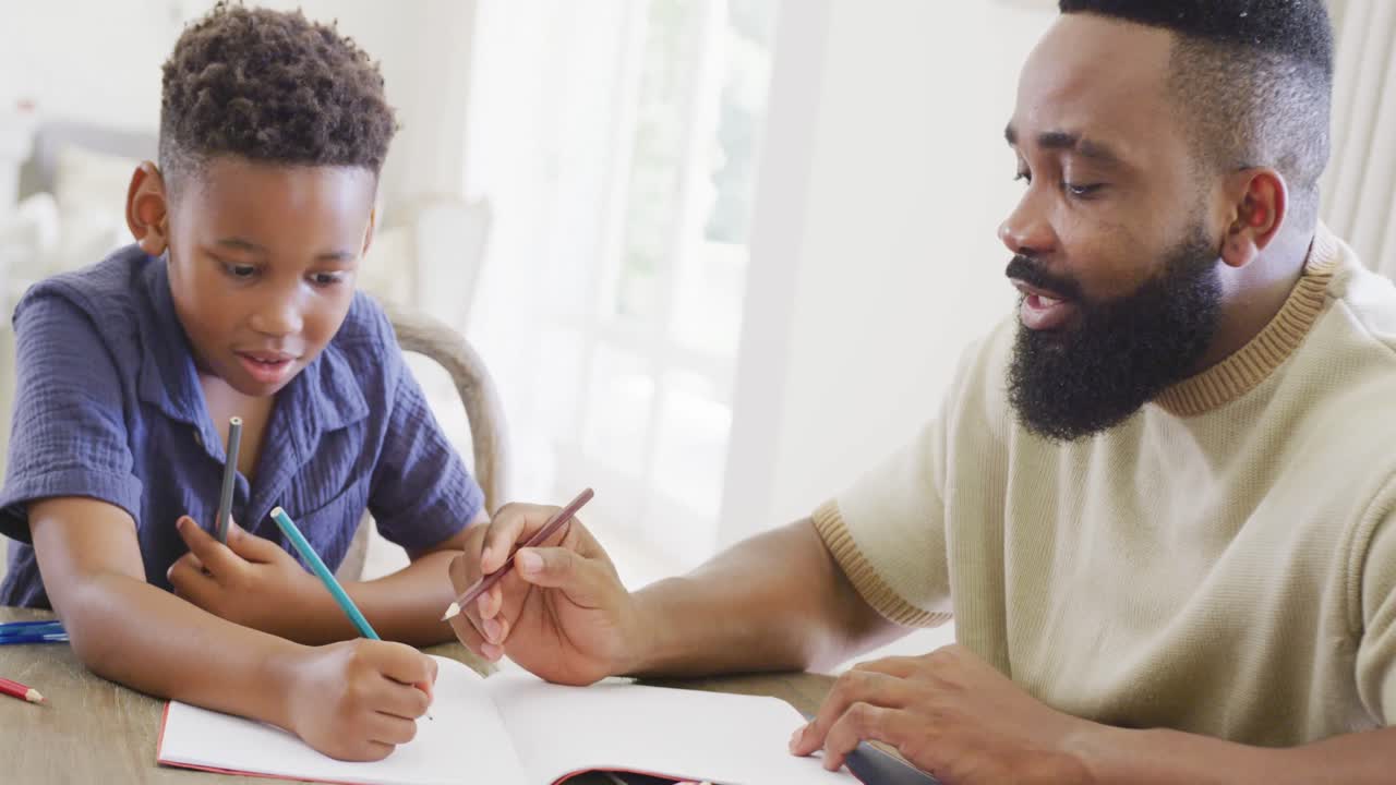 Happy african american father and son sitting at table and doing homework together, in slow motion