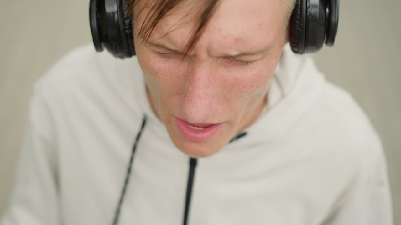 hombre blanco joven con auriculares, sudadera con capucha en el exterior, agarrando una diadema, cantando con emotividad en un ensayo, gestos expresivos, ceño fruncido, práctica de ritmo, llovizna, pelo mojado, primer plano del pavimento, intensidad, determinación