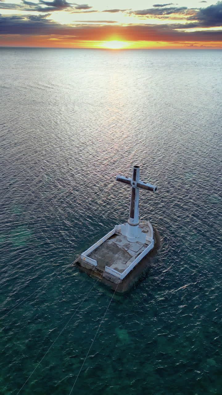 Vista aerea con drone dell'iconico monumento della Croce del Cimitero Sommerso al largo della costa dell'isola di Camiguin, Filippine. Una grande croce bianca si erge su una piattaforma sommersa nell'oceano al tramonto, a indicare un sito storico di eruzione vulcanica nel Mindanao settentrionale. Verticale.