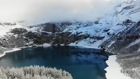 Aerial-flyover-over-the-snow-covered-trees-on-the-shores-of-lake-Oeschinensee-in-Kandersteg,-Switzerland-on-a-winter-day-with-peaks-hidden-in-clouds