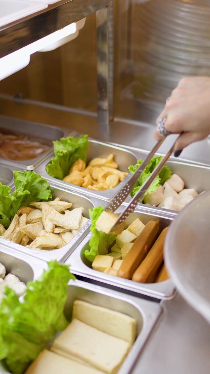 Una mano usa le pinze per prendere ingredienti crudi da una stazione di cibo a buffet hotpot asiatico ben illuminata