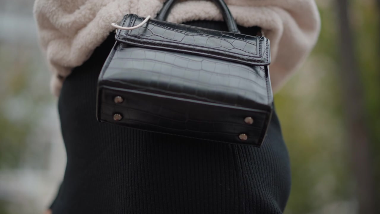 Primo piano dal basso cattura una borsa in pelle nera tenuta accanto a un maglione peloso e una gonna a costine, la borsa sollevata in avanti con un'oscillazione visibile, accessori lucidi, trama cucita e luce diurna soffusa su sfondo sfocato