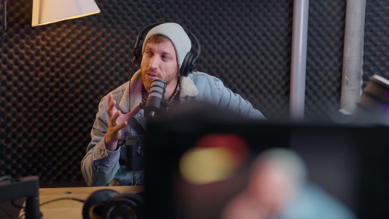 Man hosting a radio or podcast live show streaming with video camera. Person talking to recording device in professional sound or audio studio or booth. Excited and motivated hipster influencer look