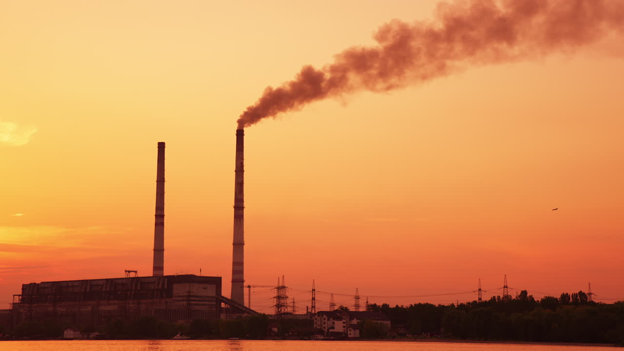 Two plant pipes towering over the plant near the river. Industrial skyline at the backdrop of beautiful orange sky.