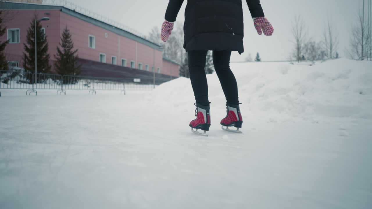 Skater gliding on outdoor ice rink during cold winter day, wearing red skates, black pants and warm coat, captured from side angle with snow-covered ground, buildings in background