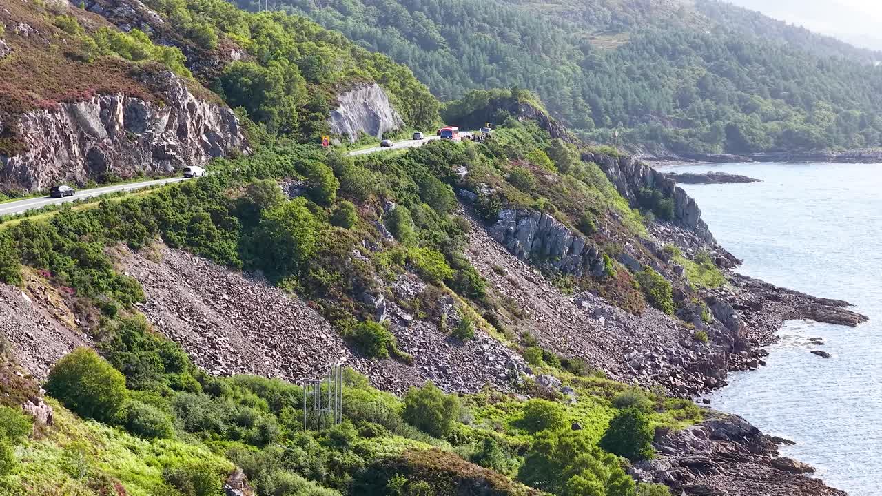 Vehicles travel on winding coastal road beside cliffs and ocean, bright daylight, smooth aerial movement