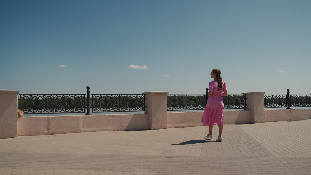 Mulher de vestido rosa caminhando no calçadão, turista parando para admirar o rio através do corrimão ornamental, calçada ensolarada e linha de árvores distante sob um céu azul claro com nuvens esparsas, tons pastel cinematográficos