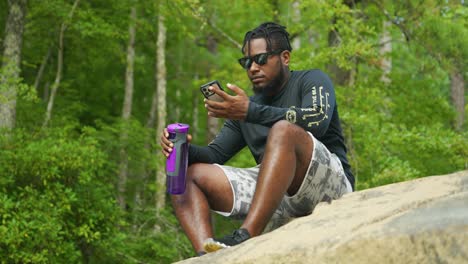 a man sits down and reads his phone on a giant rock in the middle of the woods