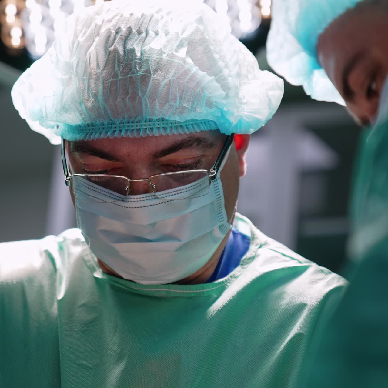 Adult male doctor in mask, cap and glasses is focused on his work. Close up portrait of a surgeon in surgery room