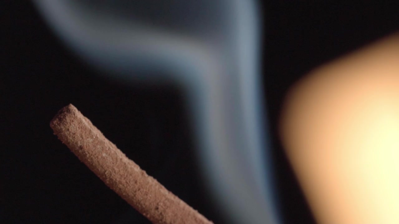 Macro view of incense stick releasing drifting smoke against dark background, highlighting aromatic ritual object, atmospheric detail, and tranquil ambiance.