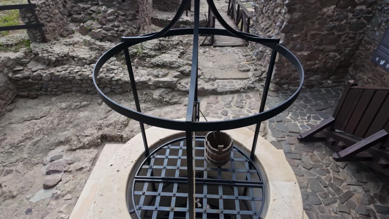 Pan right across a traditional stone well with a wooden bucket and metal frame, nestled in the historical village of Boldogkőváralja, Hungary, near castle ruins and scenic pathways