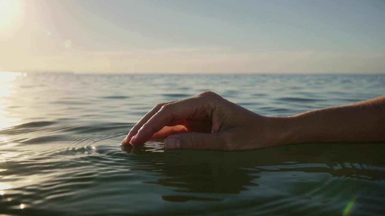 Touching adult bare forearm and hand initiating ripples on calm sea near horizon, reflecting sun