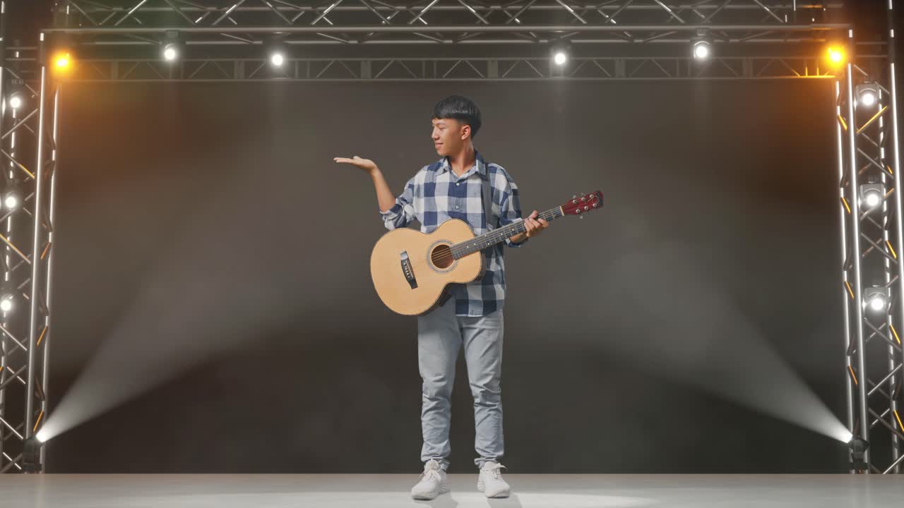 cuerpo lleno de adolescente asiático con guitarra sonriendo y señalando al lado en el escenario