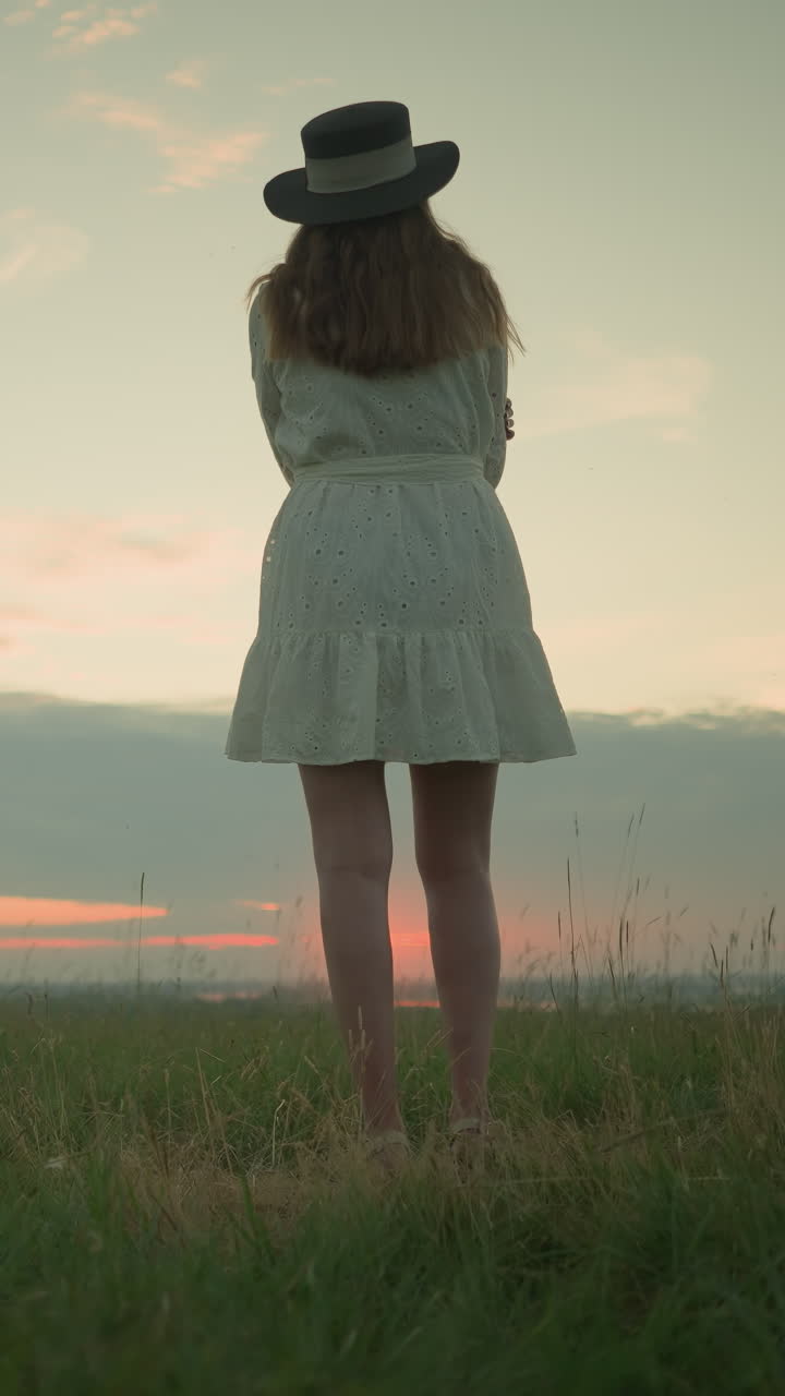 색 드레스와 모자를 입고, 팔을 접고, 조용한 잔디에서 혼자 서서, 해가 지는 동안 조용한 호수를 바라보는 여자의 뒷면.