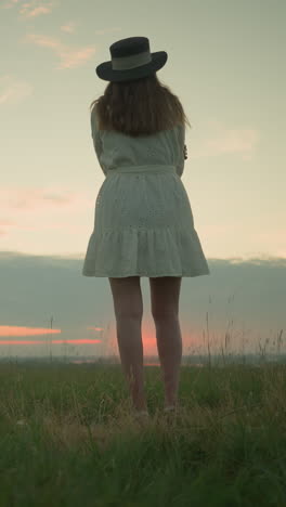 back view of a woman in a white dress and hat, arms folded, standing alone in a serene grassy field, gazing out over a tranquil lake during sunset. as the camera zooms in