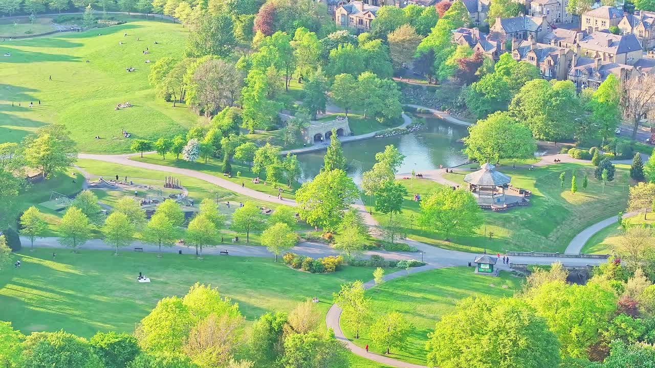 Aerial shot over a public park in Huddersfield, England, showing a curved path network, small lake with bridge, spring foliage, and an octagonal pavilion near residential buildings