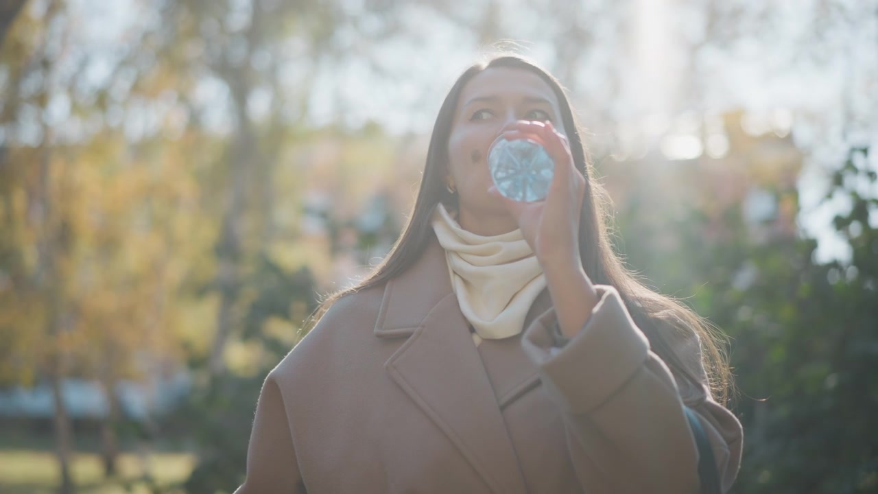 Close-up van een vrouw die een plastic waterfles opent en eruit drinkt in een park in de buitenlucht. De scène toont haar lange haar, bruine jas en crèmekleurige sjaal, terwijl de vrouw de dop met één hand losdraait.