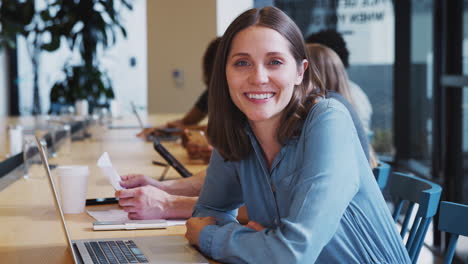 Portrait-Of-Businesswoman-Working-At-Desk-On-Laptop-In-Shared-Open-Plan-Office-Workspace