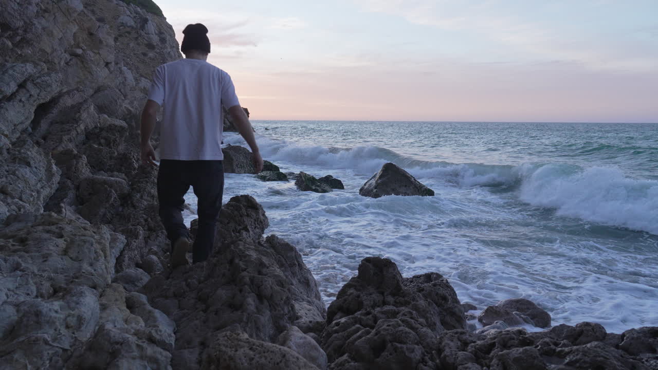 el hombre camina sobre rocas cerca del mar mientras las olas aplastan la costa, se pone en cuclillas para disfrutar, toma una foto, mientras la luz de la mañana es demasiado hermosa