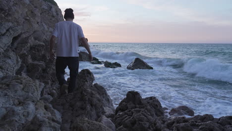 der mann geht auf felsen in der nähe des meeres, während die wellen an land brechen, er hockt sich hin, um zu genießen, macht ein foto, während das morgenlicht zu schön ist