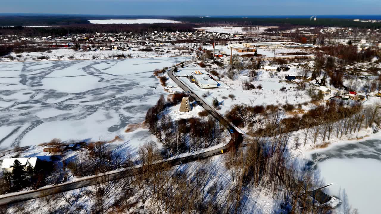 A frozen lake with winding ice patterns and a nearby small town surrounded by snow-covered fields under clear winter skies, captured from an elevated aerial view.