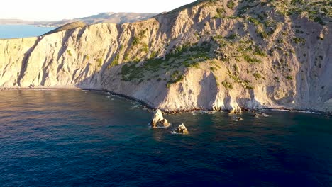 panning aerial drone shot of deep blue waves crashing against the cliffs of a greek peninsula during golden hour sunrise or sunset