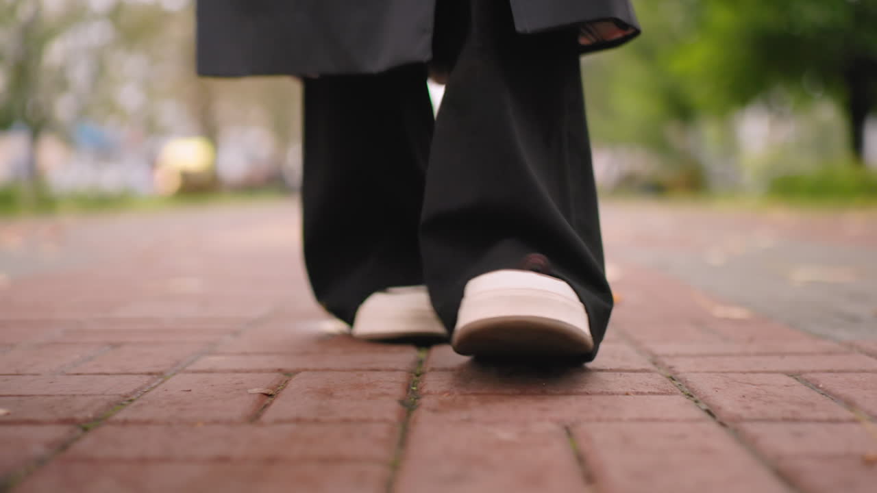 Close-up of woman walking on brick path wearing black pants and white sneakers, autumn leaves scattered on ground, capturing step motion, casual urban lifestyle concept with seasonal atmosphere outdoors