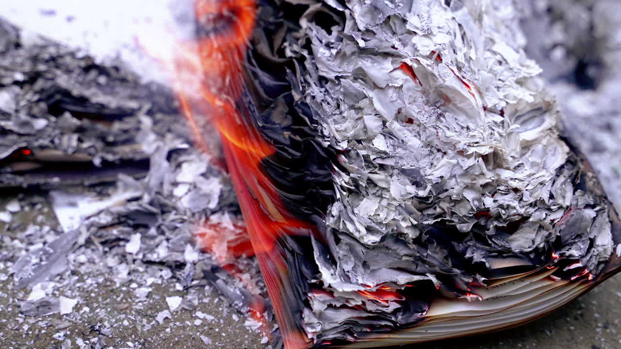 Libro aperto che prende fuoco su una superficie di pietra, circondato da cumuli di cenere di carta grigia e braci. Libro aperto che brucia a terra con cenere grigia.