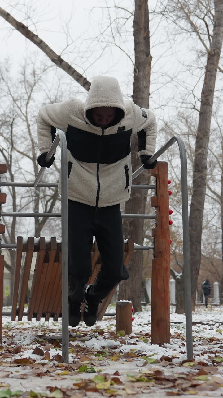 joven adulto realiza un intenso entrenamiento de construcción muscular en barras paralelas en un parque urbano nevado, vestido con una sudadera cálida y guantes, se levanta en un ejercicio de inmersión mientras la gente camina en la distancia