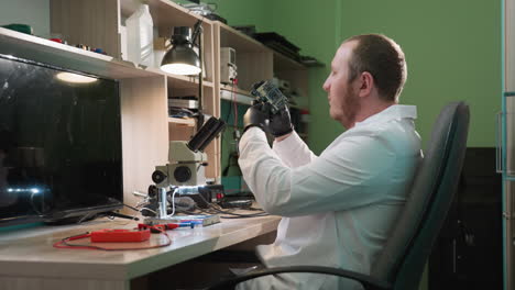 una vista de cerca de un técnico en una bata blanca de laboratorio inspeccionando una placa de circuito. el técnico examina la placa de circuito bajo un microscopio con varios equipos en los estantes