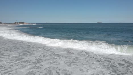 Backwards-aerial-movement-following-the-incoming-rough-long-waves-coming-in-on-Ipanema-beach-with-the-Arpoador-rock-and-islands-in-the-background-on-a-sunny-day-in-Rio-de-Janeiro