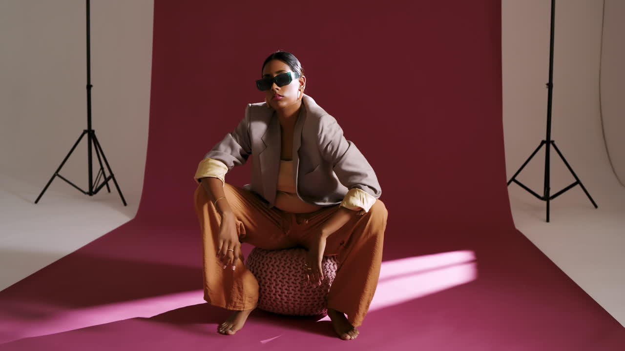 Noticing light pattern, Indian woman in beige blazer adjusting sunglasses, posing on pouf in studio