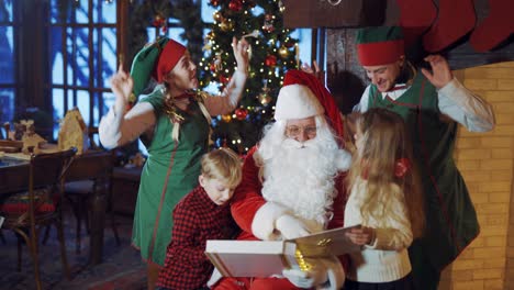 santa with little kids and elves dancing on christmas background. santa claus is showing photo album to children while joyful elves having fun inside the house.