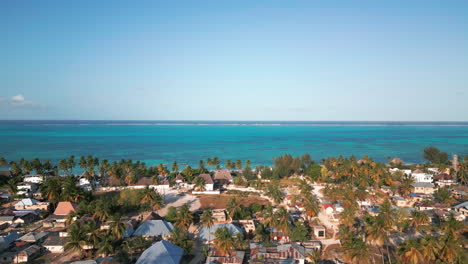 A-tropical-coastal-village-with-palm-trees-and-thatched-roof-structures,-bordering-a-serene-turquoise-ocean-under-a-clear-blue-sky