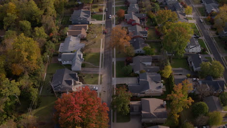 Sobrevuele-Las-Casas-Suburbanas-Y-Las-Hojas-De-Otoño-Mientras-Un-Automóvil-Blanco-Conduce-Por-La-Calle-Y-Se-Sale-Del-Marco