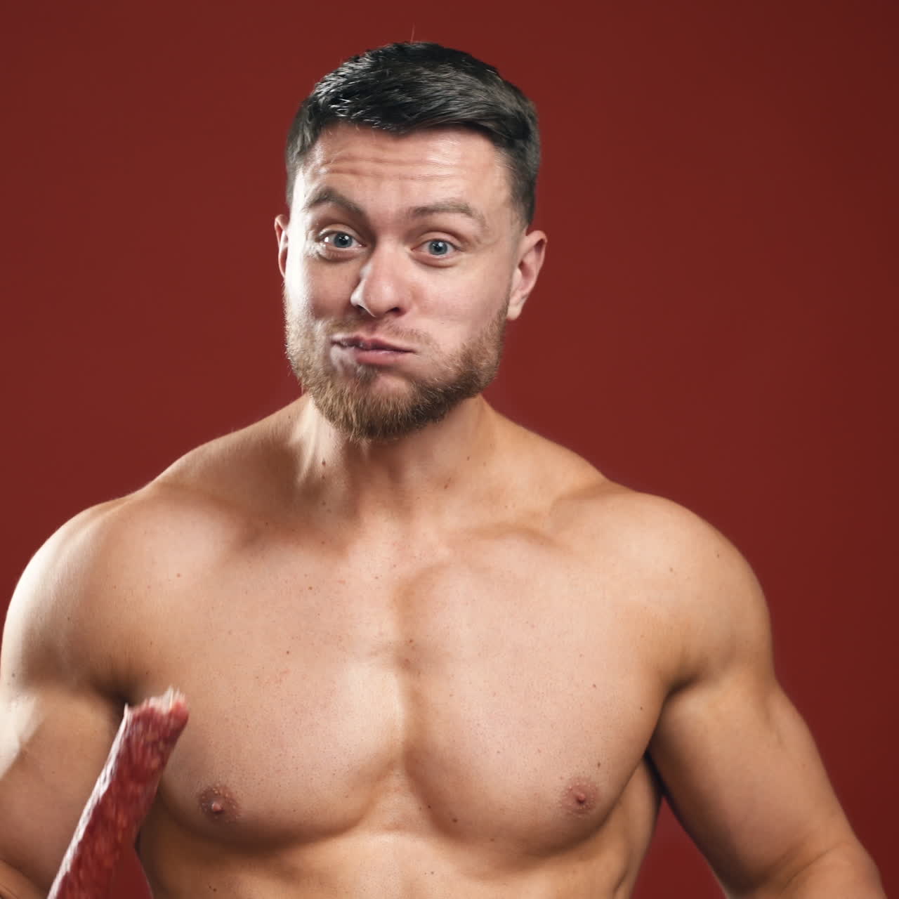 Hungry strong male eating unhealthy food. Half-length portrait of muscular man without shirt eats bread and sausage quickly on the studio background.