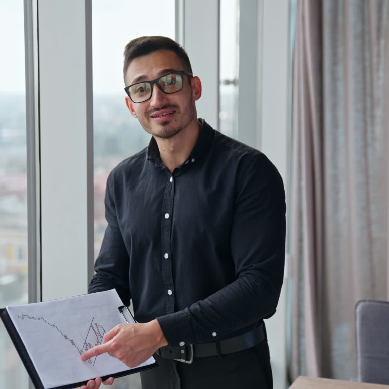 Businessman in black shirt, trousers and glasses holding a paper with chart. Man following the graph, explaining it to camera
