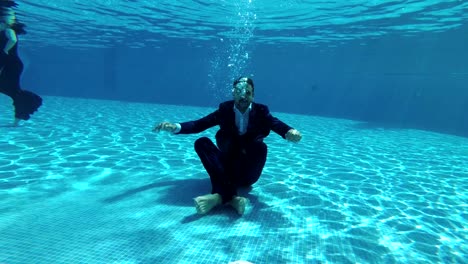 the happy groom in a suit and a white shirt goes down underwater to the bottom of the pool. he sits at the bottom in a lotus position, looks at the camera and smiles. slow motion. action camera underwater.