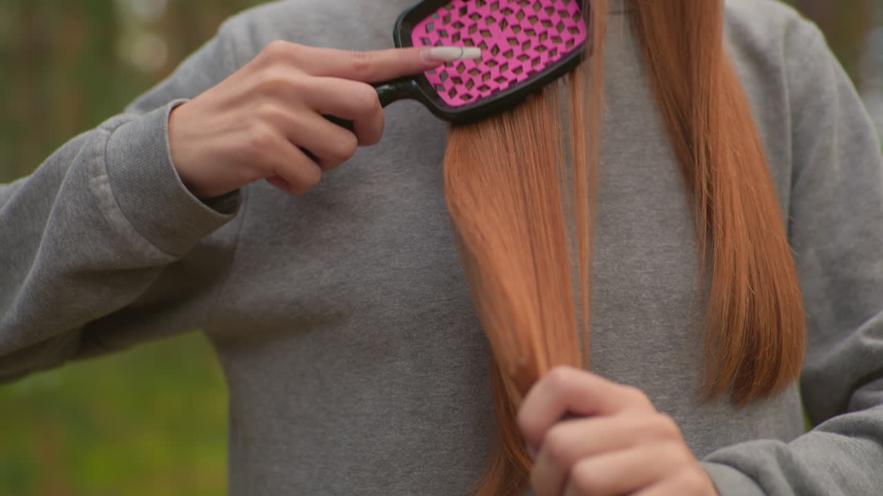 Close-up of young woman brushing her straight, reddish-brown hair with a pink hairbrush against her gray sweater in a serene forest setting