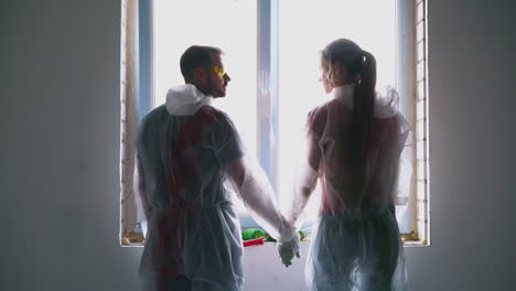 positive couple in protective wear joins hands near window