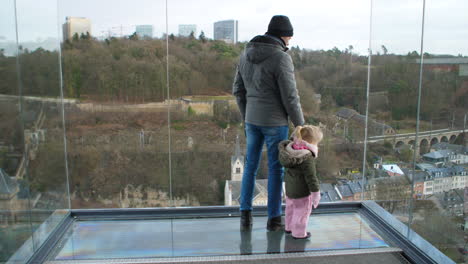 wide shot of father - young daughter overlooking old city of luxembourg from glass platform during winter.