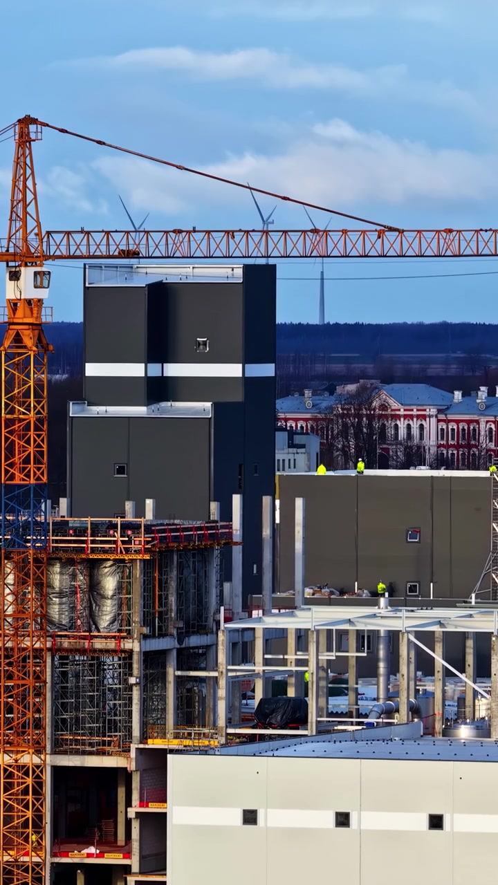 Close-up drone flight over a large industrial construction site in a rural town in Northern Europe, with wind turbines in the background, showcasing a cold industrial complex.